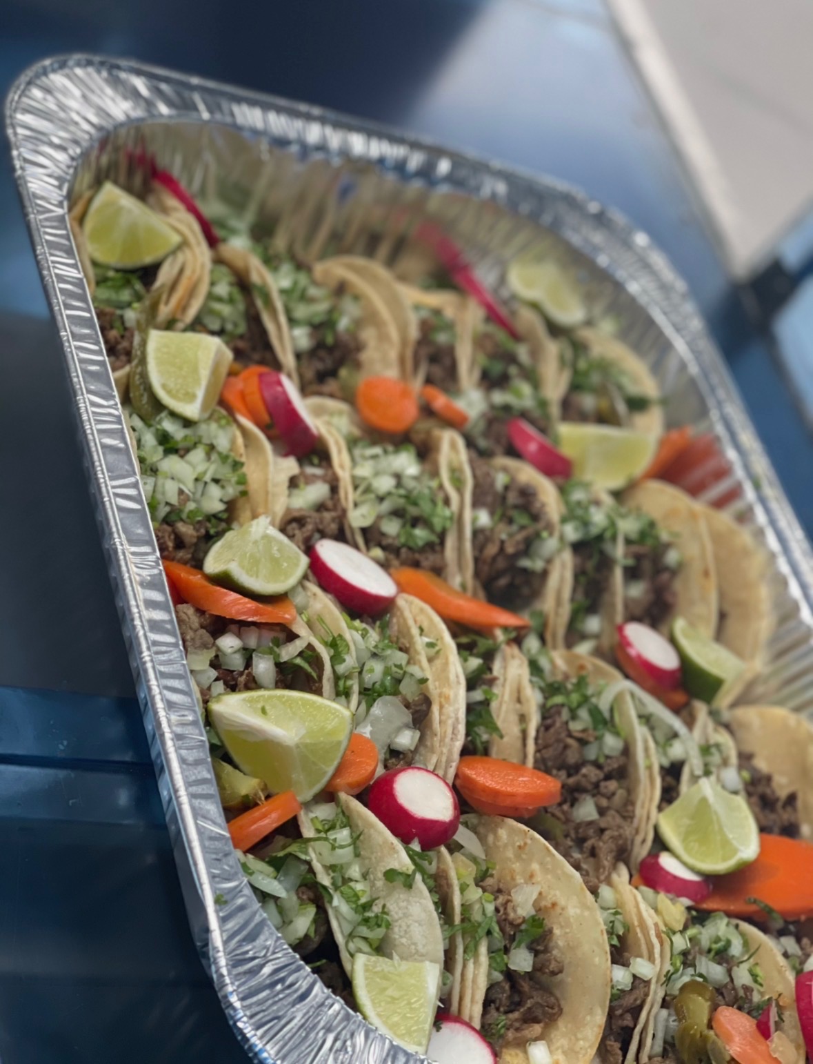 Aluminum tray filled with street tacos topped with onion, cilantro, radish, and lime